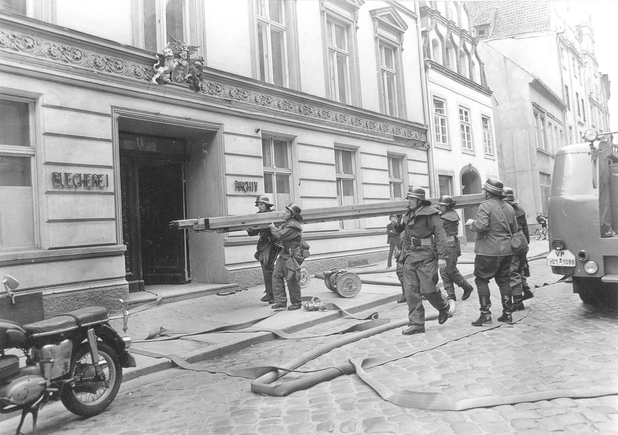 Das Bild zeigt mehrere Feuerwehrmänner aus dem Jahre 1974, die vor der Stadtbibliothek eine Leiter tragen.