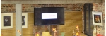 Ein Fernseher zeigt das Logo der Stadtbibliothek während Kinder ihre Hände heben und ein Schild mit einem kleinen Gesicht während einer Buchvorstellunghochhalten