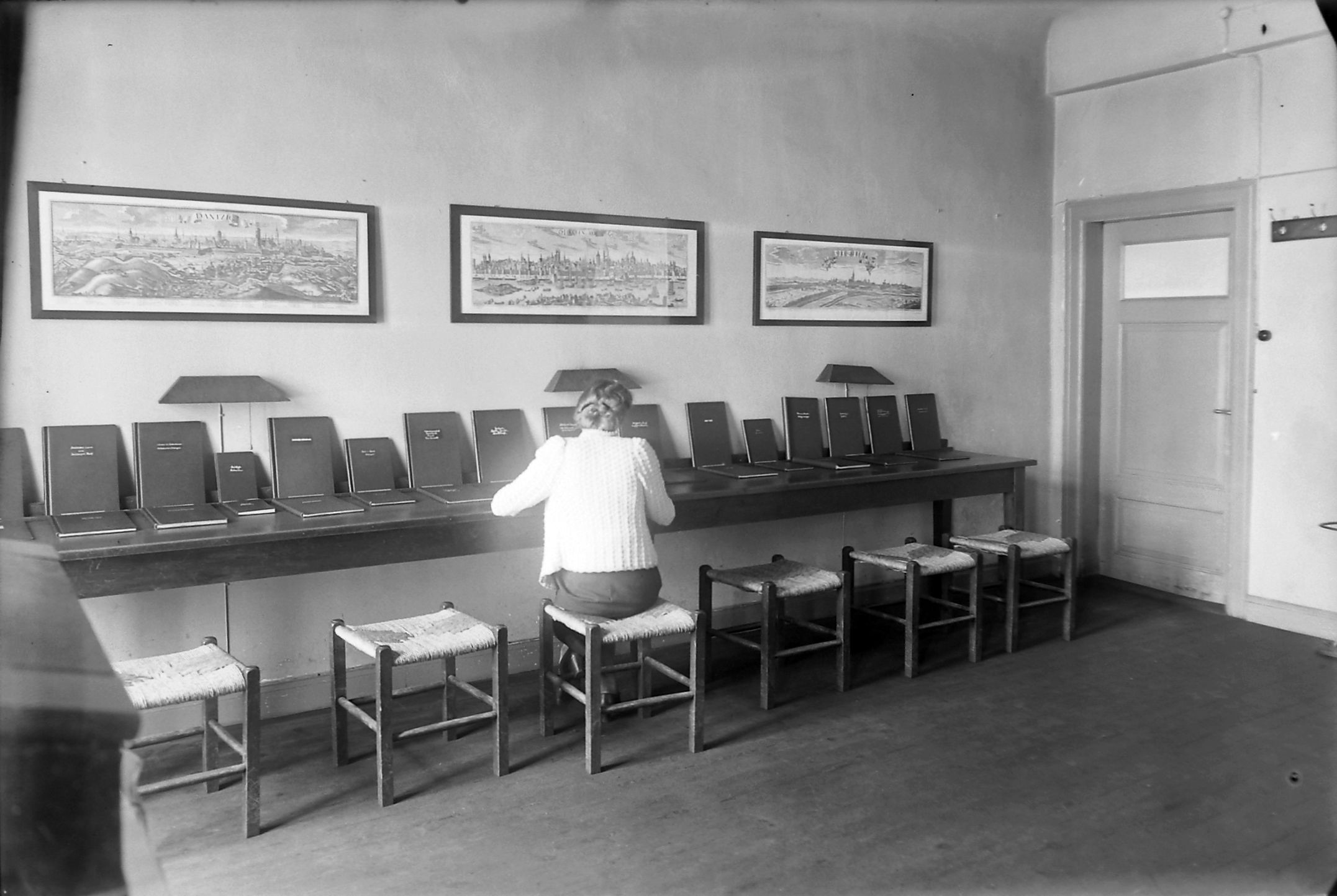 Eine Frau sitzt an einem großen Tisch auf dem viele Bücher ausgelegt sind im Jahr 1938.