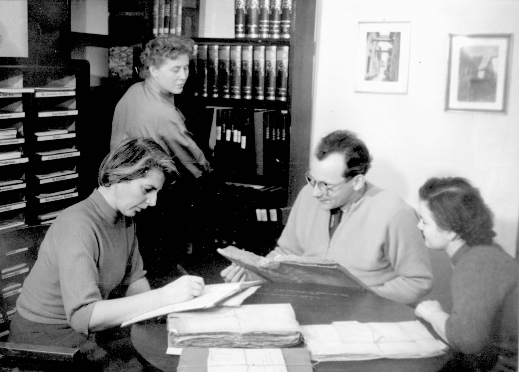 Drei Personen sitzen an einem Tisch und schauen in Bücher im Jahr 1965.