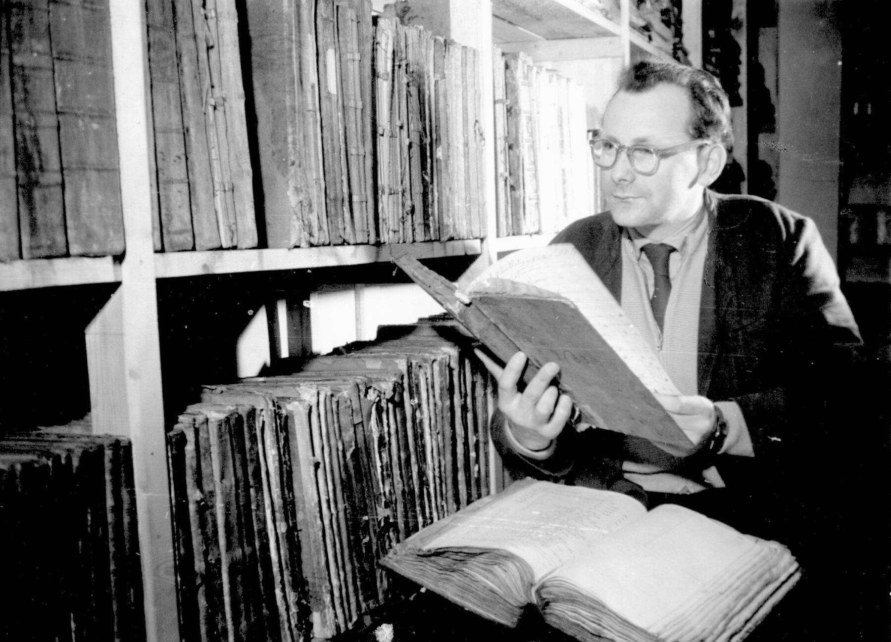 Ein Mann sitz vor einem Bücherregal mit einem Buch auf seinen Beinen und hält ein altes Buch in der Hand