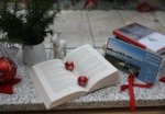 Ein geöffnetes Buch liegt auf einem Tisch mit einer weißen Tischdecke. Auf dem Buch liegen rote Christbaumkugeln. In einer Vase stehen Tannenzweige. Rechts steht ein weiterer Bücherstapel