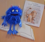 Ein blaues Monster sitzt auf einem Buch und grinst. Daneben liegt ein Buch mit dem Titel 